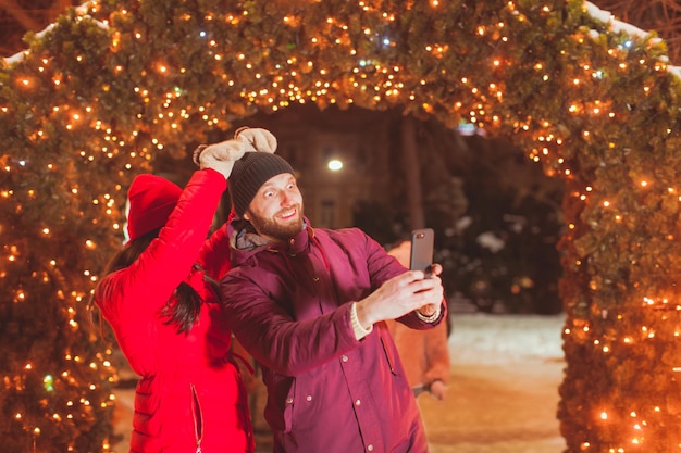 Joven hombre y mujer feliz haciendo selfie con caras graciosas de pie cerca del arco con decoración navideña, al aire libre en el mercado de invierno