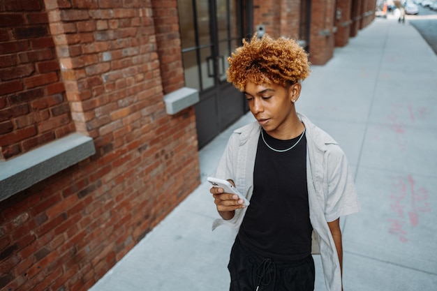 Joven hombre multirracial positivo con ropa elegante sosteniendo un teléfono móvil inteligente mientras camina