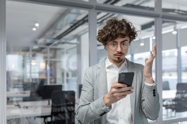 Un joven hombre de negocios adulto de cabello rizado parece confundido mientras usa un teléfono móvil en una oficina moderna