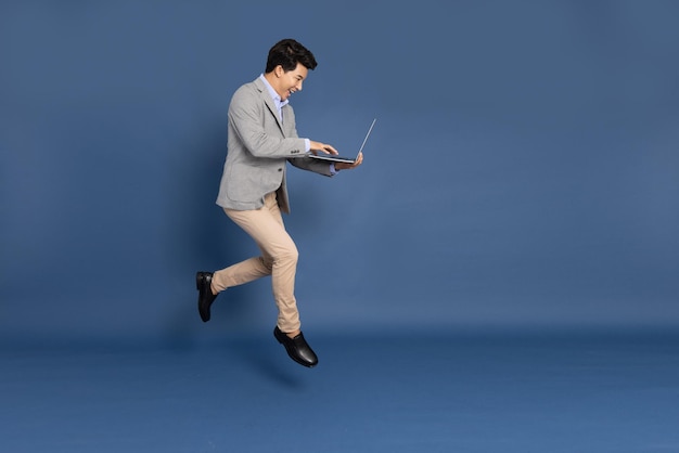 Joven hombre de negocios asiático saltando en el aire usando una computadora portátil aislada en un fondo azul profundo