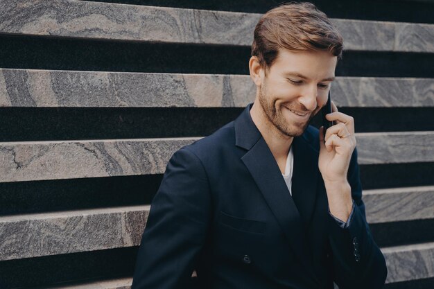 Joven hombre de negocios atractivo y confiado con barba ordenada en ropa formal hablando por teléfono móvil al aire libre