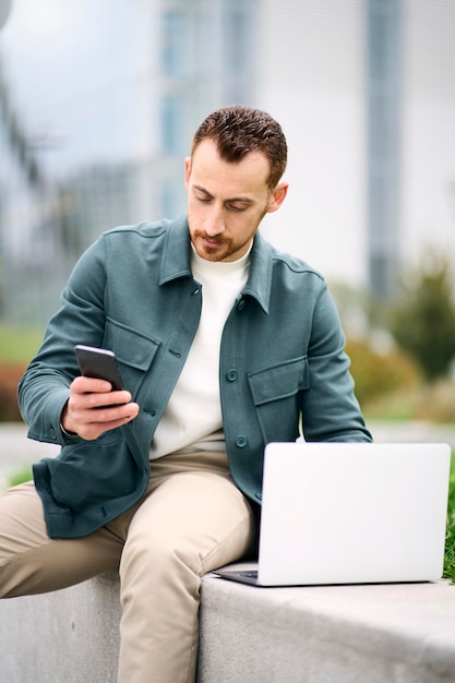 Joven hombre de negocios atractivo revisando el mensaje del teléfono inteligente mientras trabaja con una computadora portátil