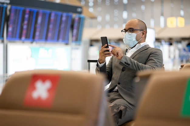 Joven hombre de negocios con bolsa de carrito usando un teléfono inteligente para registrarse en vuelo. Viajar durante la pandemia debe ser distanciamiento social. Él sentado en la terminal del aeropuerto.