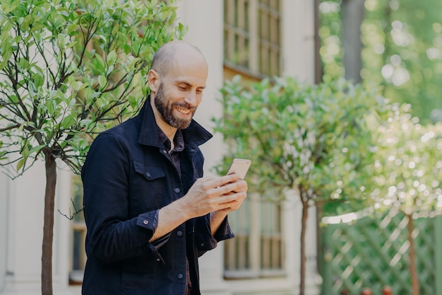 Un joven hombre de negocios calvo positivo con una espesa barba oscura feliz de recibir un mensaje de felicitación celebra poses de cumpleaños contra una hermosa vista al parque Concepto de personas y tecnologías modernas