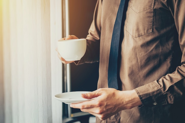 Joven hombre de negocios la celebración de taza de café en la oficina en la ventana.