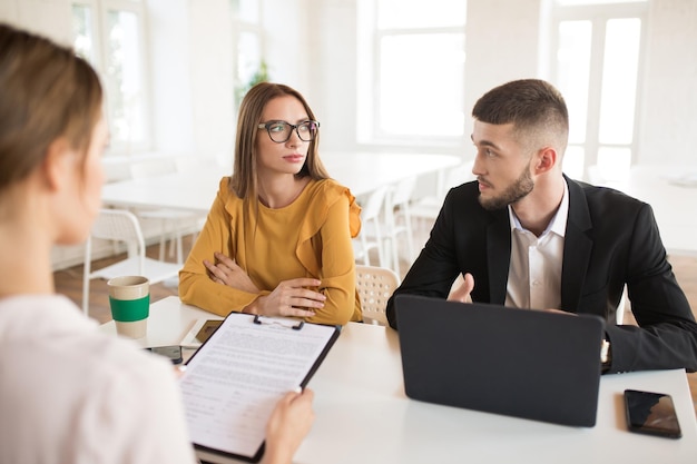 Joven hombre de negocios con computadora portátil y mujer de negocios con anteojos mirándose cuidadosamente mientras habla con el solicitante sobre el trabajo Jóvenes empleadores pasan una entrevista de trabajo en una oficina moderna