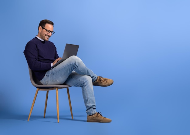 Joven hombre de negocios confiado con gafas usando una computadora portátil mientras está sentado en una silla sobre un fondo azul