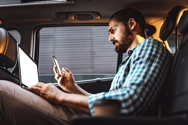 Un joven hombre de negocios confiado sostiene una computadora portátil y un teléfono inteligente en la mano y envía mensajes de texto en el asiento trasero del auto.