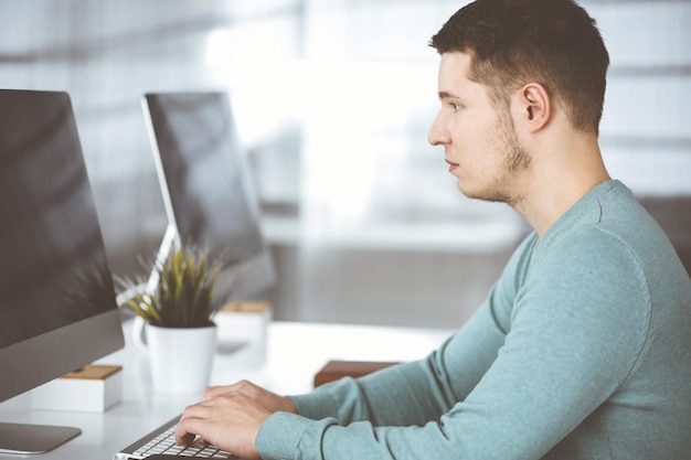 Un joven hombre de negocios confiado, vestido con un suéter verde, está sentado en el escritorio de una oficina moderna, mientras trabaja en la computadora en su nuevo proyecto. Foto de cabeza de empresario masculino o especialista en TI en wo