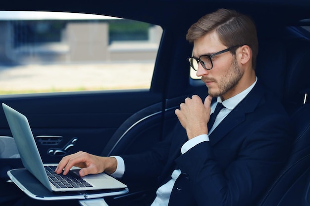 Un joven hombre de negocios considerado con la mano en la barbilla mientras se sienta en el auto de lujo y usa su computadora portátil