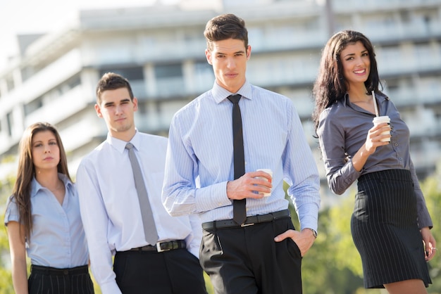 Joven hombre de negocios, elegantemente vestido, de pie con su equipo en un descanso para tomar café frente al edificio de oficinas y mirando a la cámara con una expresión seria en su rostro.