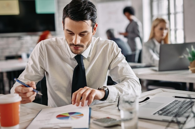 Un joven hombre de negocios está leyendo datos y analizando el progreso del negocio mientras revisa el papeleo en la oficina Hay personas en el fondo