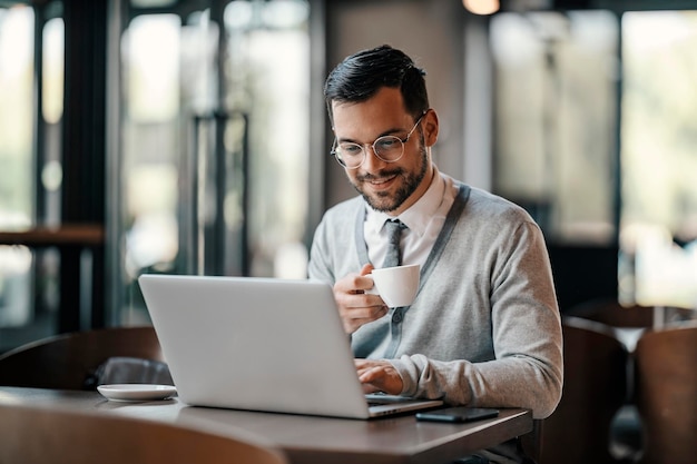 Un joven hombre de negocios feliz en causal inteligente está sentado en un café tomando café y escribiendo en una computadora portátil