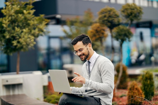 Un joven hombre de negocios feliz con un estilo casual inteligente está sentado afuera en el parque y escribiendo en una computadora portátil