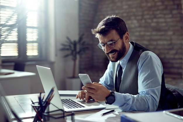 Joven hombre de negocios feliz que usa un teléfono inteligente mientras trabaja en una computadora portátil en la oficina