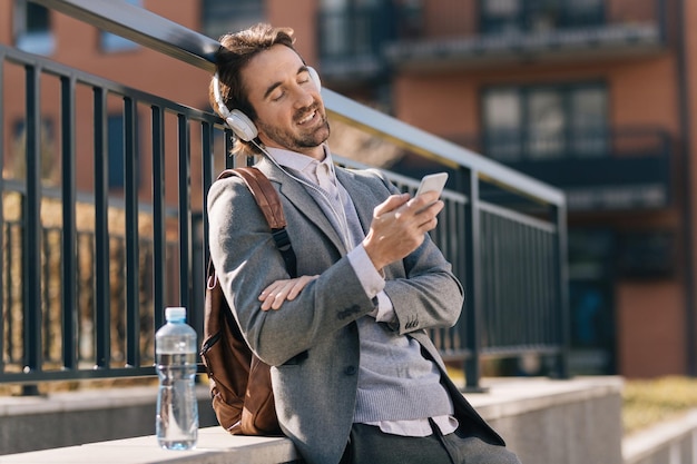 Joven hombre de negocios feliz usando teléfono móvil mientras escucha música con auriculares en la ciudad