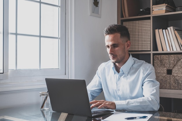 Joven hombre de negocios millennial caucásico feliz trabajando en casa en el escritorio con computadora portátil o computadora divirtiéndose Joven hombre en videoconferencia cuidando del negocio o mercado