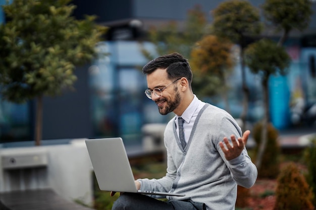 Un joven hombre de negocios de moda está sentado al aire libre y teniendo una conferencia telefónica en la computadora portátil