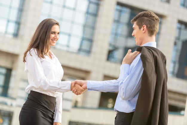 Foto joven hombre de negocios y mujer de negocios, elegantemente vestidos, estrechando la mano frente a la empresa mirándose el uno al otro sonriendo feliz cooperación comercial.