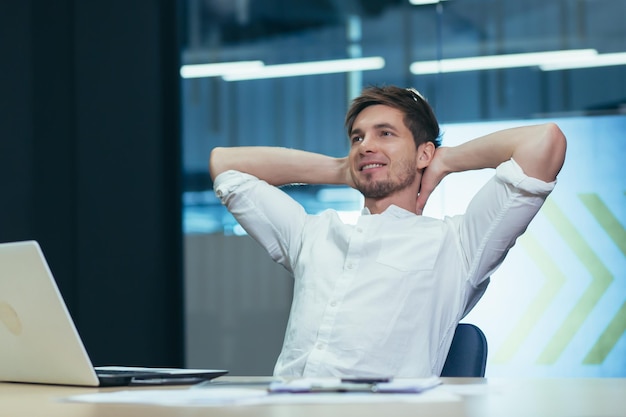 Un joven hombre de negocios en la oficina está descansando y soñando tirando sus manos detrás de su cabeza un hombre en el trabajo trabajando con una computadora portátil