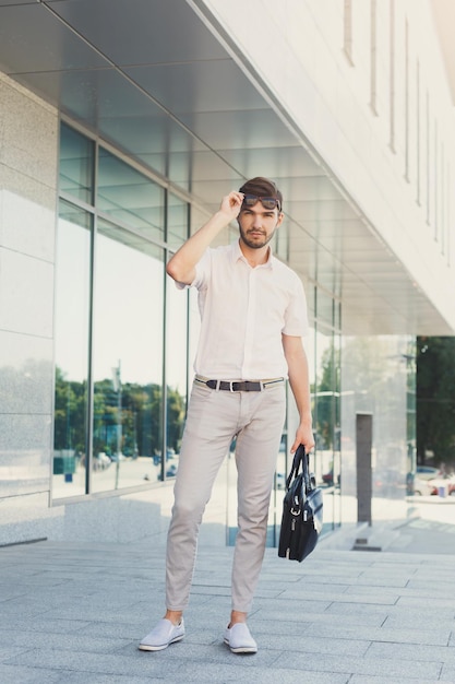 Joven hombre de negocios pensativo quitándose las gafas de sol y mirando atentamente mientras sostenía el maletín, parado cerca del moderno edificio de oficinas, de cuerpo entero, copiando espacio. Concepto de negocio y éxito