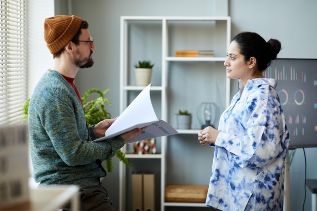 Joven hombre de negocios serio con manual hablando con una colega en ropa informal inteligente y respondiendo