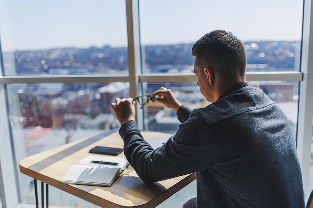 Un joven hombre de negocios se sienta en una mesa y sostiene anteojos para la corrección de la visión en sus manos Descanse después del trabajo en una computadora portátil