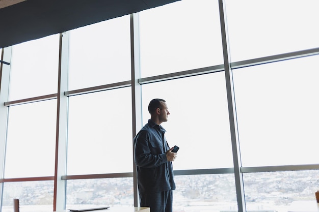 Joven hombre de negocios sosteniendo un teléfono en una oficina o centro de negocios en el contexto de grandes ventanales de un rascacielos Concepto de trabajo y carrera