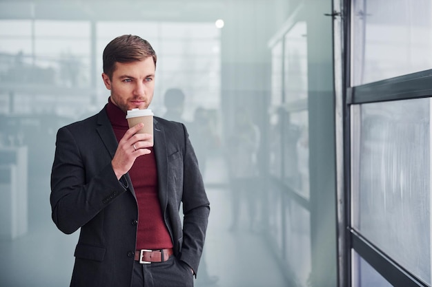 Un joven hombre de negocios con traje de lujo y ropa formal con una taza de bebida está en el interior de la oficina.