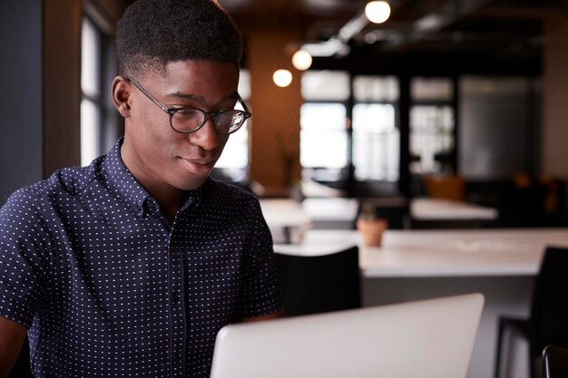 Joven hombre negro creativo sentado en un escritorio en una oficina de planta abierta usando una computadora portátil vista lateral de cerca