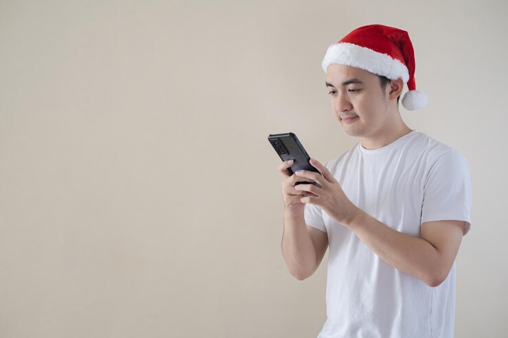 El joven hombre de Papá Noel asiático sostiene y mira un teléfono inteligente en un fondo beige aislado