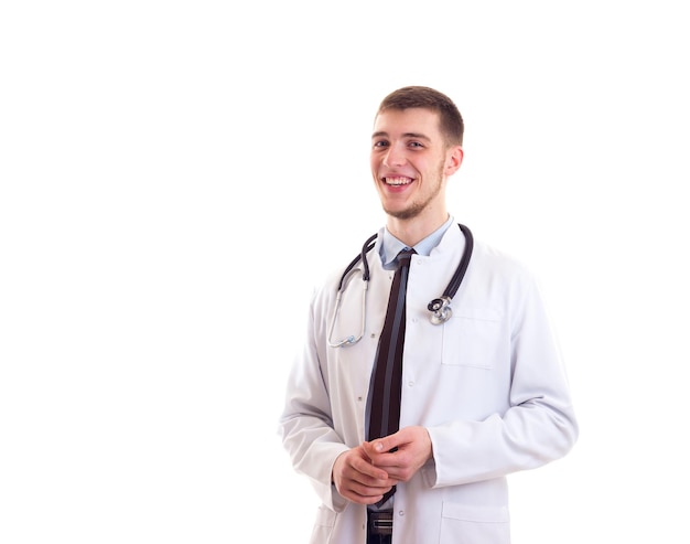Joven hombre positivo con cabello castaño con corbata de camisa azul y bata de médico sobre fondo blanco en el estudio
