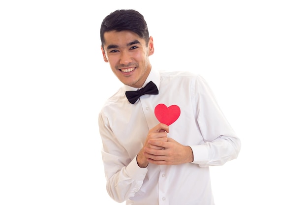 Joven hombre positivo con cabello oscuro en camiseta blanca con pajarita negra sosteniendo un corazón de papel rojo sobre fondo blanco en el estudio