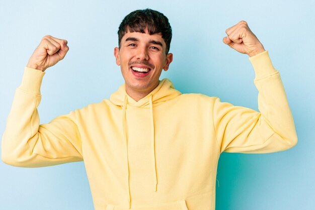 Joven hombre de raza mixta aislado de fondo azul que muestra un gesto de fuerza con los brazos, símbolo del poder femenino