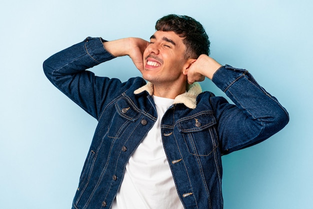 Joven hombre de raza mixta aislado de fondo azul sintiéndose confiado, con las manos detrás de la cabeza.