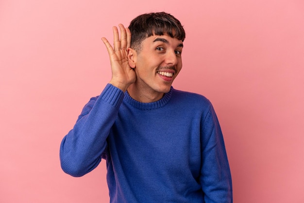 Joven hombre de raza mixta aislado de fondo rosa tratando de escuchar un chisme