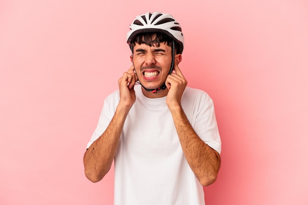 Joven hombre de raza mixta con una bicicleta de casco aislado en un fondo rosado cubriendo las orejas con las manos.