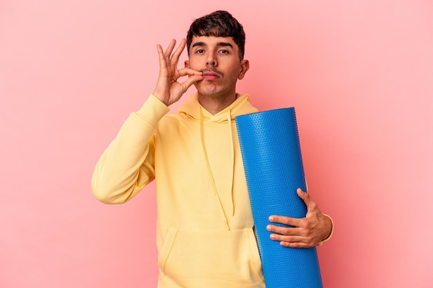 Joven hombre de raza mixta sosteniendo una alfombra deportiva aislada en un fondo rosado con los dedos en los labios guardando un secreto.