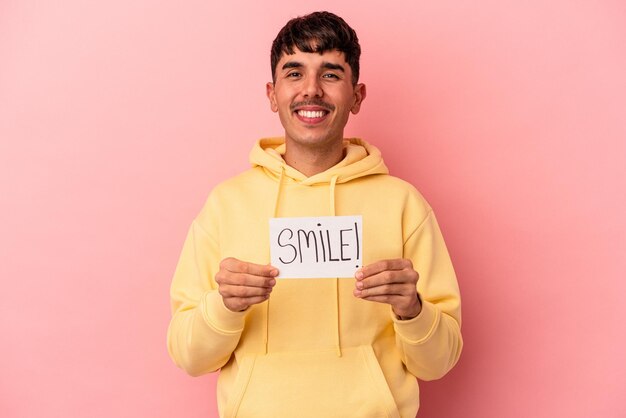 Joven hombre de raza mixta sosteniendo un cartel de sonrisa aislado sobre fondo rosa