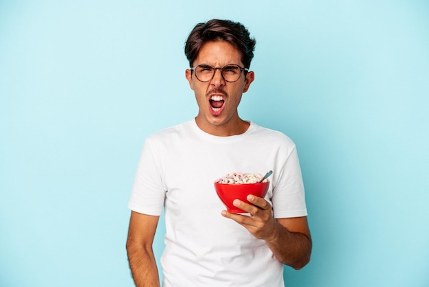 Joven hombre de raza mixta sosteniendo cereales aislados de fondo azul gritando muy enojado y agresivo.