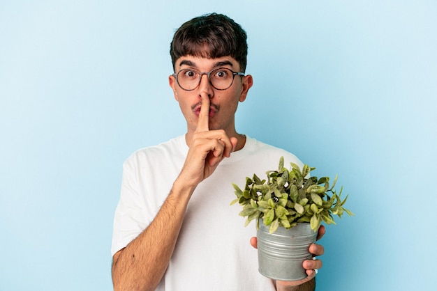 Joven hombre de raza mixta sosteniendo una planta aislada de fondo azul guardando un secreto o pidiendo silencio.