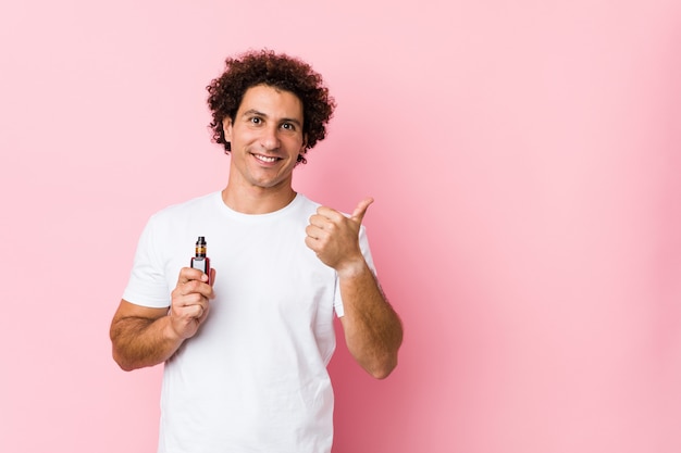 Joven hombre rizado caucásico sosteniendo un vape sonriendo y levantando el pulgar