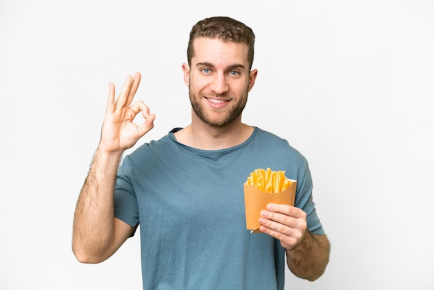 Joven hombre rubio guapo sosteniendo papas fritas sobre fondo blanco aislado que muestra el signo de ok con los dedos