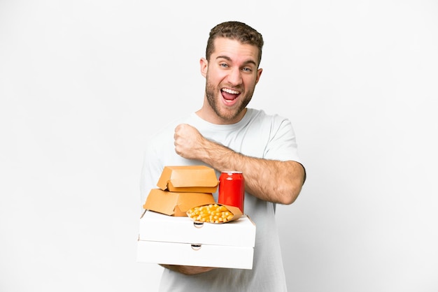 Joven hombre rubio guapo sosteniendo pizzas y hamburguesas sobre un fondo aislado celebrando una victoria