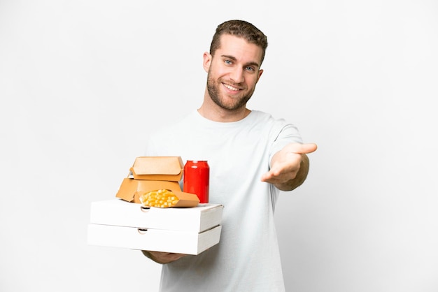 Joven hombre rubio guapo sosteniendo pizzas y hamburguesas sobre un fondo aislado dándose la mano para cerrar un buen trato