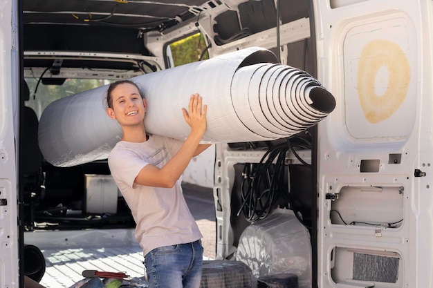 Joven hombre sonriente feliz que lleva un rollo de goma de aislamiento de agujeros en el hombro con una furgoneta convertida en el fondo