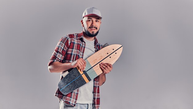 Joven inconformista con una camisa a cuadros y una gorra con un longboard sobre un fondo gris.