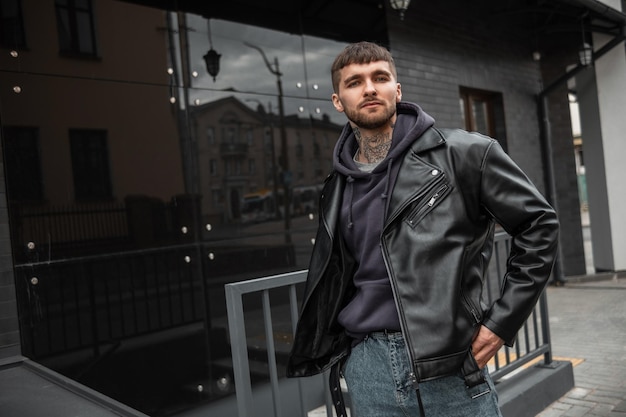 Un joven inconformista con estilo, con pelo, barba y un tatuaje en el cuello, vestido con ropa informal de moda, con una chaqueta de cuero con capucha y jeans, camina cerca de un edificio moderno negro