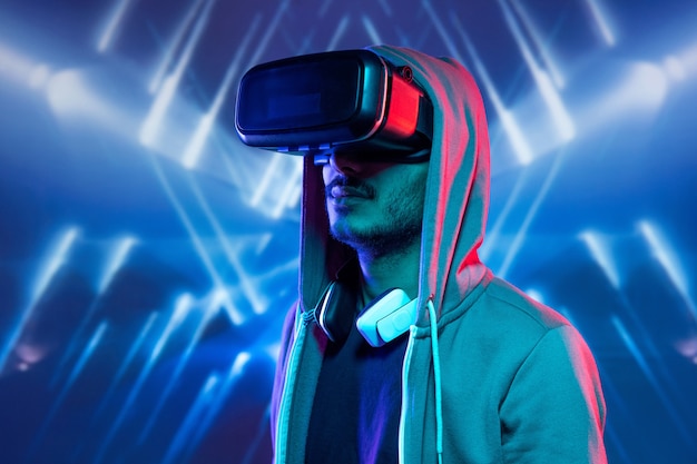 Joven inconformista con rastrojo inmerso en la realidad virtual con sudadera con capucha viendo películas en 3D contra el fondo azul luminoso