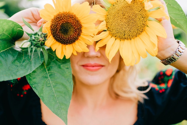 Joven increíble hermosa rubia en vestido negro con retrato de estilo de vida de patrón étnico. Moda country. Linda extraña feliz alegre mujer en ropa tradicional eslava posando en girasoles al aire libre